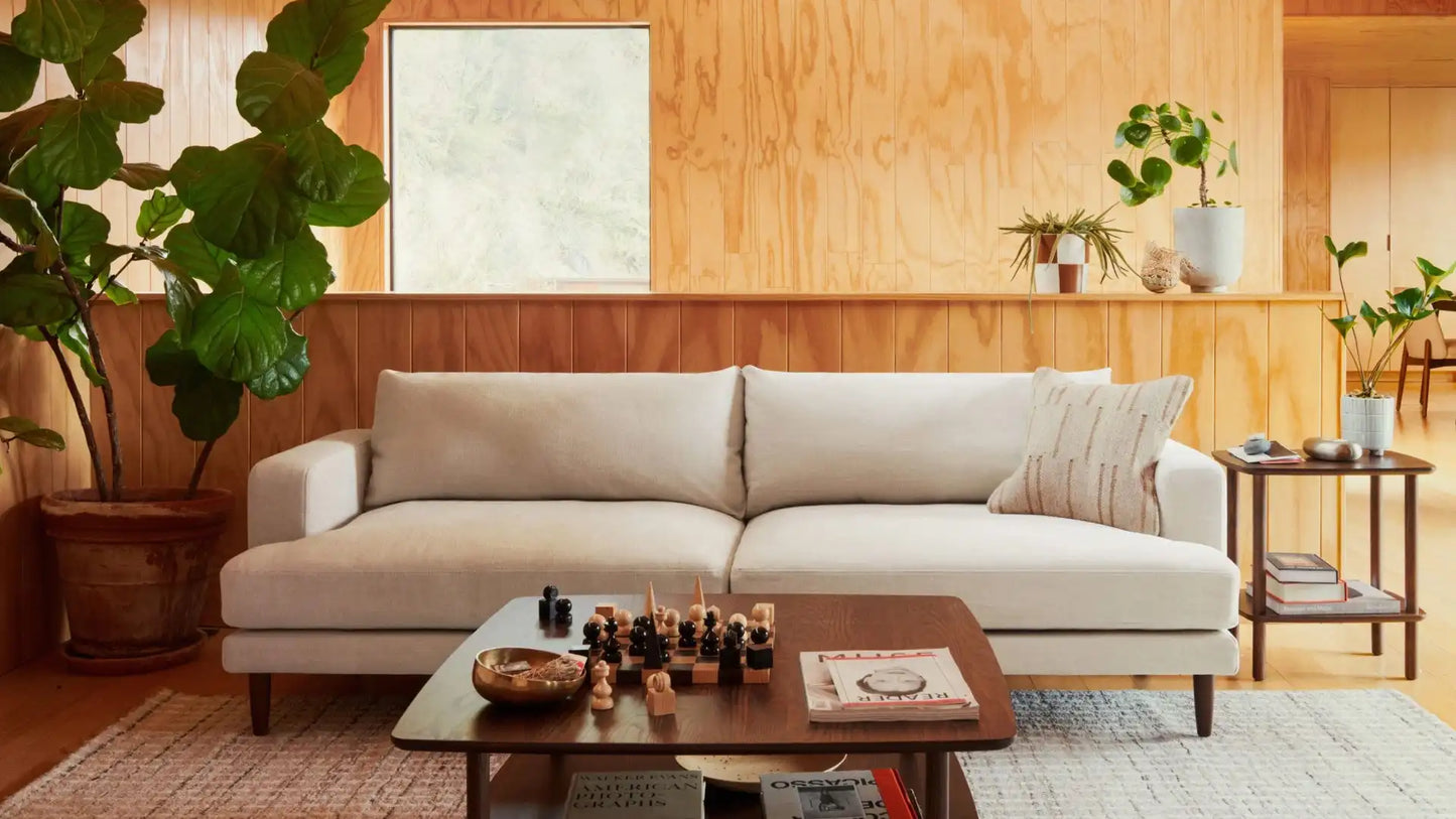 A cozy living room with an Ember Mid-Century Modern 75" Sofa in light beige, a wooden coffee table adorned with chess pieces, and lush green plants. The space is enhanced by wooden walls and a large window, creating a warm, inviting atmosphere.