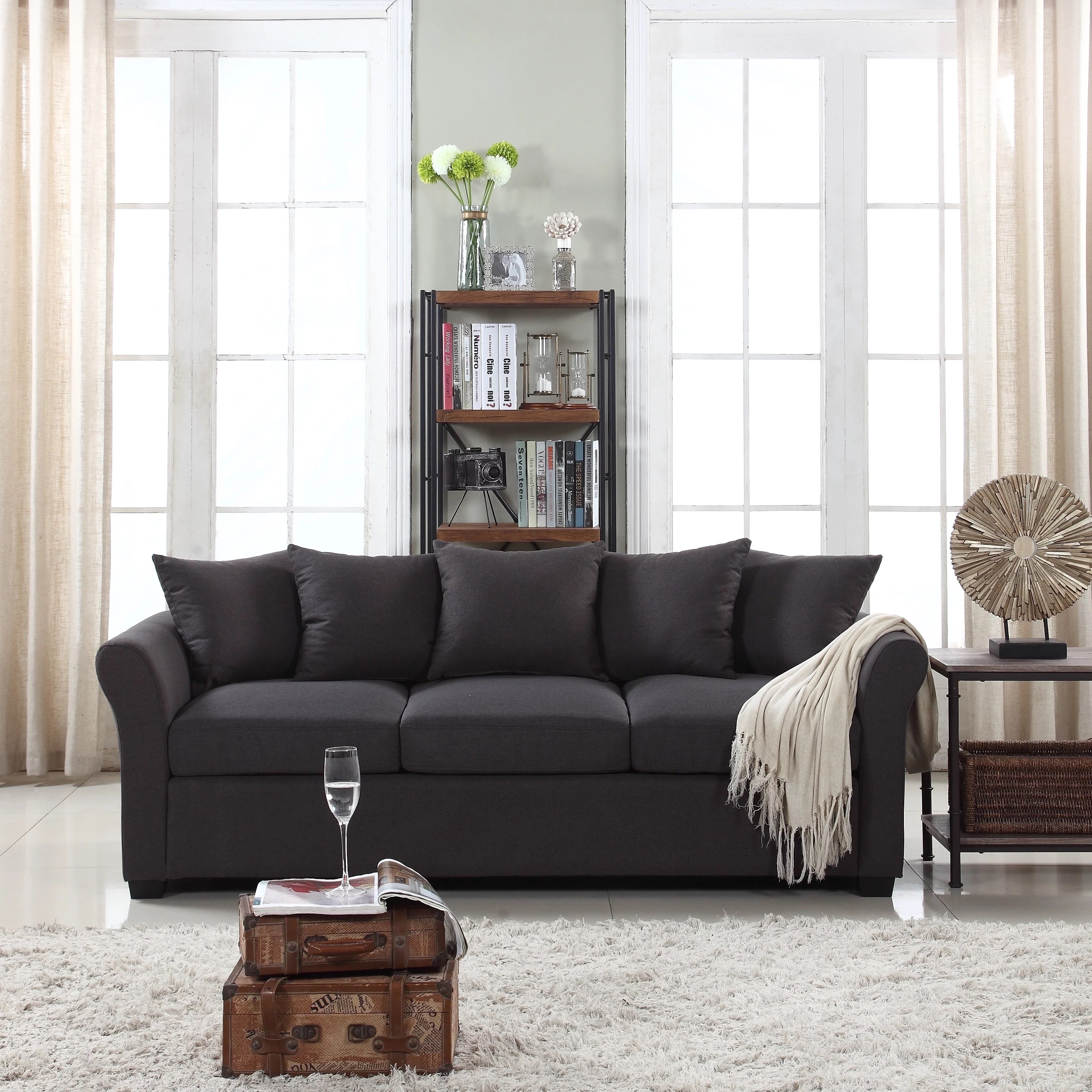 A cozy living room featuring a dark grey sofa with four cushions and a light throw blanket on the right arm. Behind the sofa is a tall black bookshelf with decor items. Large windows with light curtains flank the scene, and a glass of water sits on a stack of vintage suitcases.