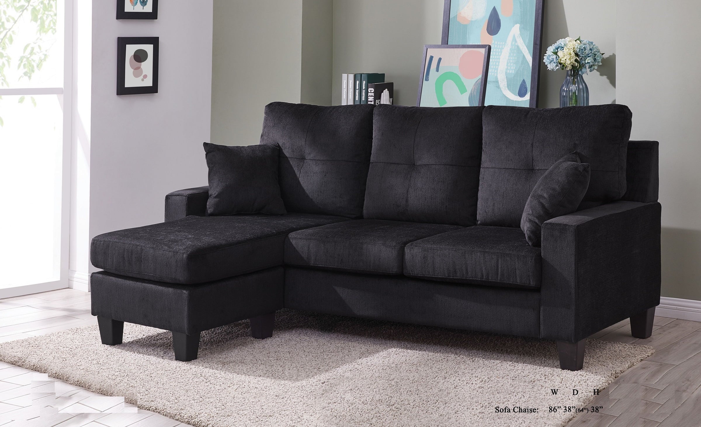 A modern black sectional sofa with an L-shaped design, featuring plush cushions and a matching chaise lounge on the left side. The sofa is placed in a brightly lit room with light-colored walls, abstract wall art, framed pictures, and a textured beige rug.