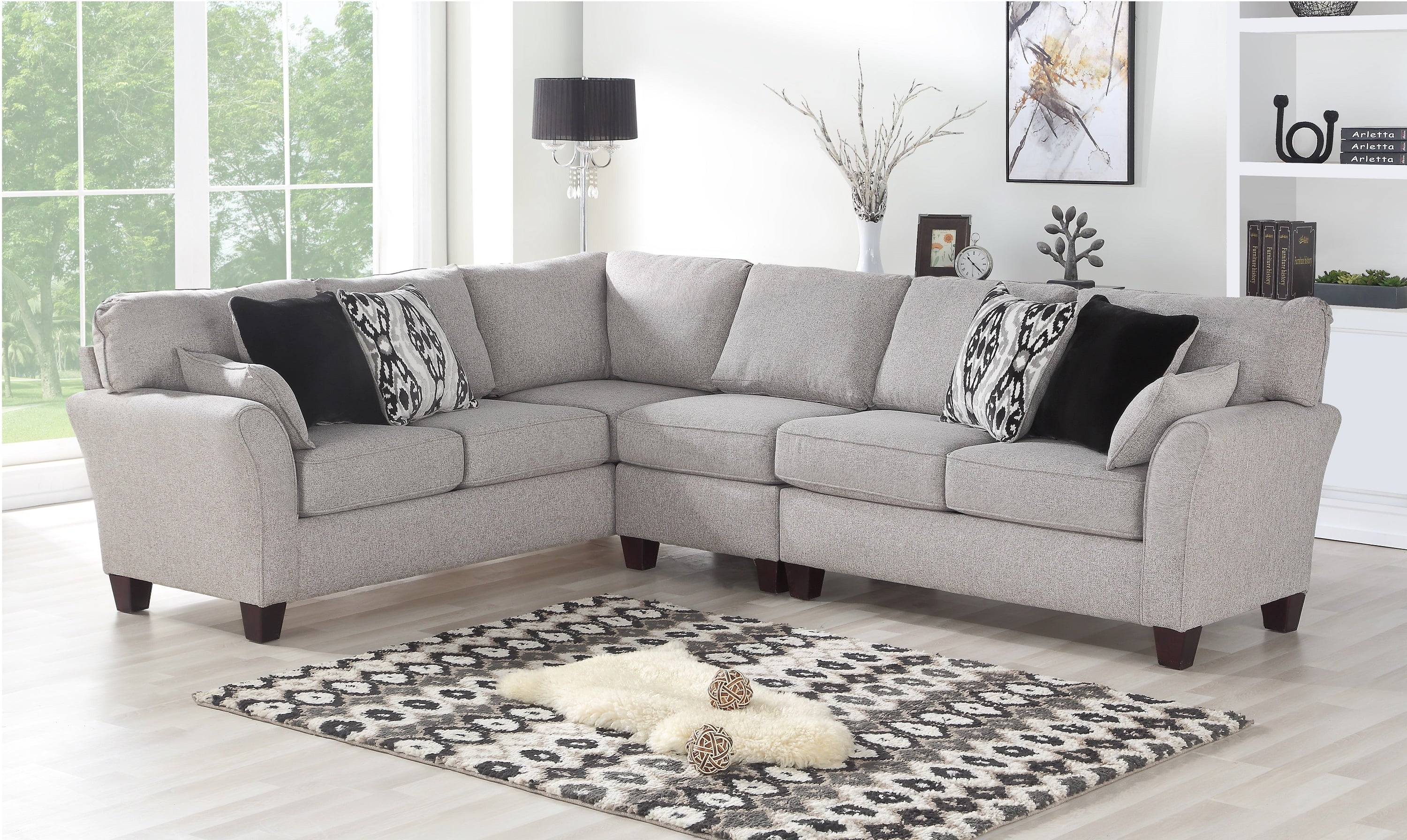 A light grey L-shaped sectional sofa with black and white patterned pillows is set in a bright, modern living room. The room features large windows, a patterned rug with a fur accent, a black lamp, a white shelf with books, and decorative items.