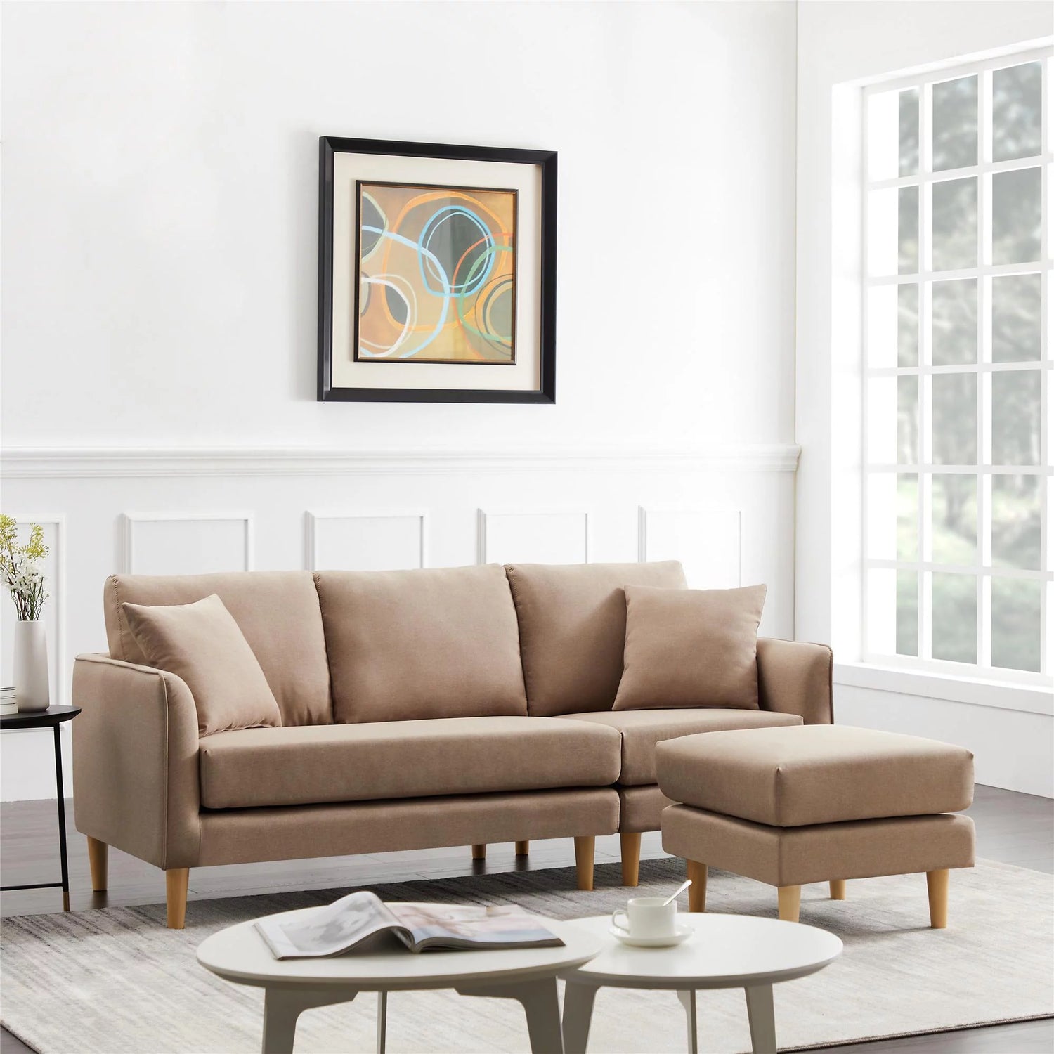 A light beige sofa with matching ottoman in a modern living room with white walls and wainscoting. Above the sofa, there's a framed abstract painting with circular patterns. A coffee table with books and a cup is in the foreground, and large windows allow natural light in.