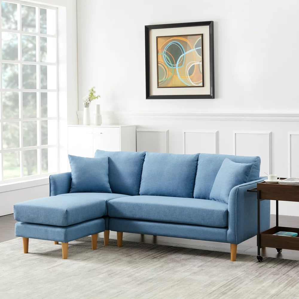 A modern living room features a light blue sectional sofa with wooden legs. A framed abstract painting hangs above the sofa. A window to the left allows natural light to fill the room, highlighting a white paneled wall and a wooden side table with decor.