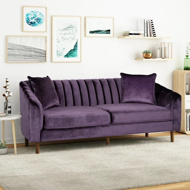 Purple velvet sofa with two matching cushions, wooden legs, situated in a modern living room. The wall behind the sofa features a gallery of abstract art in white frames. The room includes a light wooden side table with a plant and books, a cabinet, and a rug.