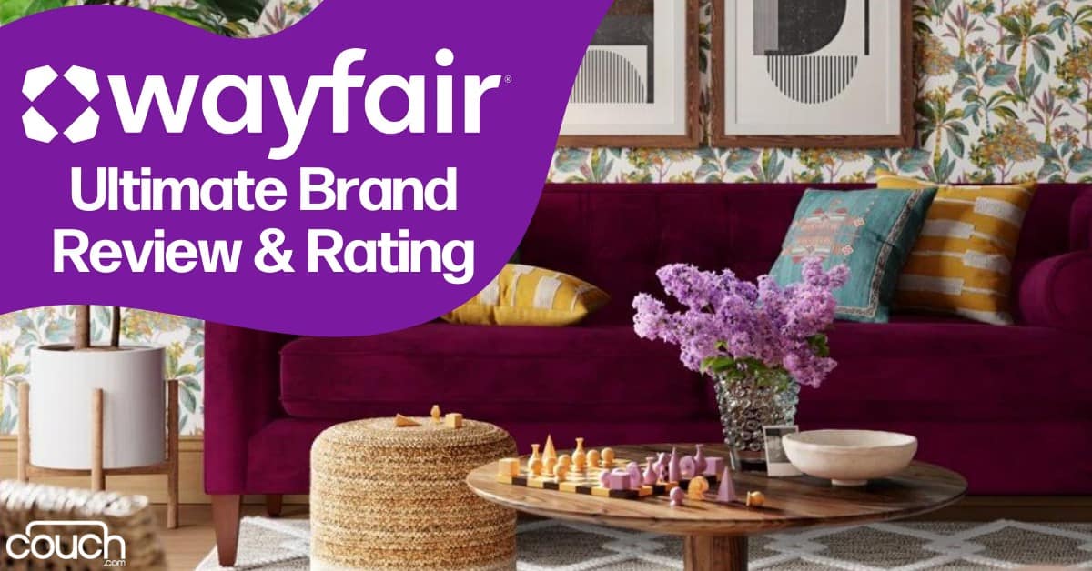 A stylish, cozy living room features a deep maroon sofa adorned with colorful throw pillows, a round wooden coffee table with a vase of purple flowers, and a woven ottoman. The wall behind the sofa has patterned wallpaper with framed art. Text reads "Wayfair Ultimate Brand Review & Rating".