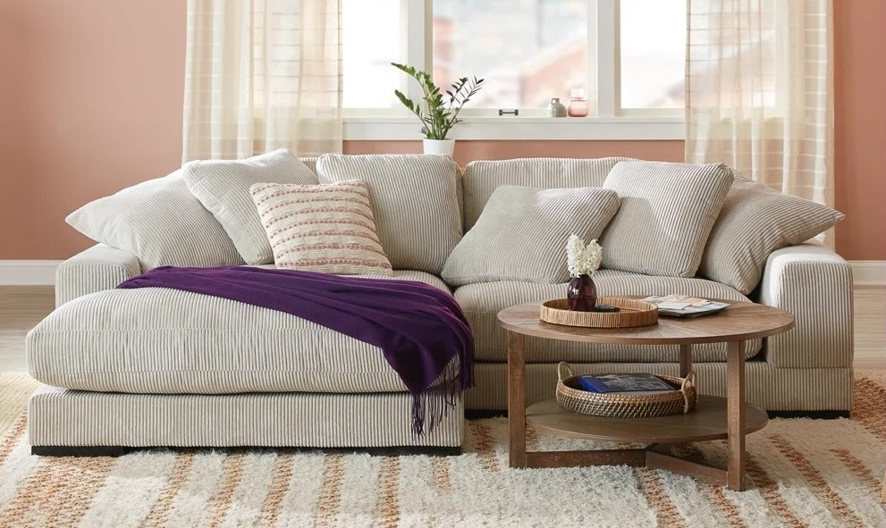 A cozy living room features a light-colored sectional sofa adorned with several pillows and a purple throw blanket. A wooden round coffee table with books and a small plant sits in front of the sofa. The room has soft peach walls and a large window with light curtains.