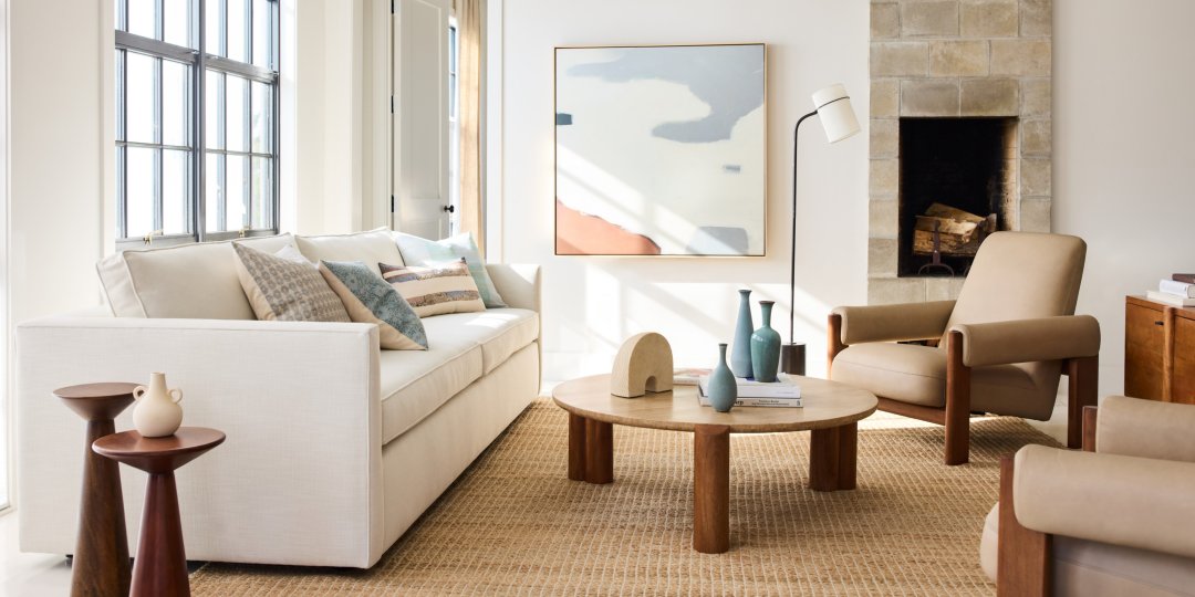 A bright, modern living room features a white sofa, two beige armchairs, and a circular wooden coffee table with decorative items like vases and a sculpture. A large abstract painting hangs above a stone fireplace, and natural light streams in through wide windows.