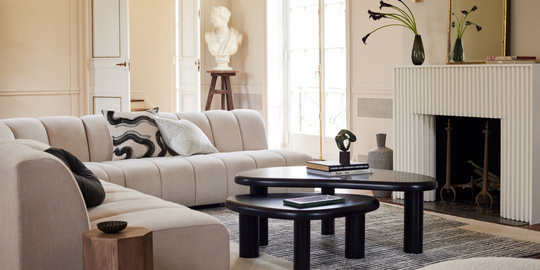 A modern living room with a curved beige sofa adorned with black and white throw pillows. There's a black coffee table on a striped rug in front of a white fireplace. Decorative items include a vase with flowers, a sculpture on a pedestal, and several books.