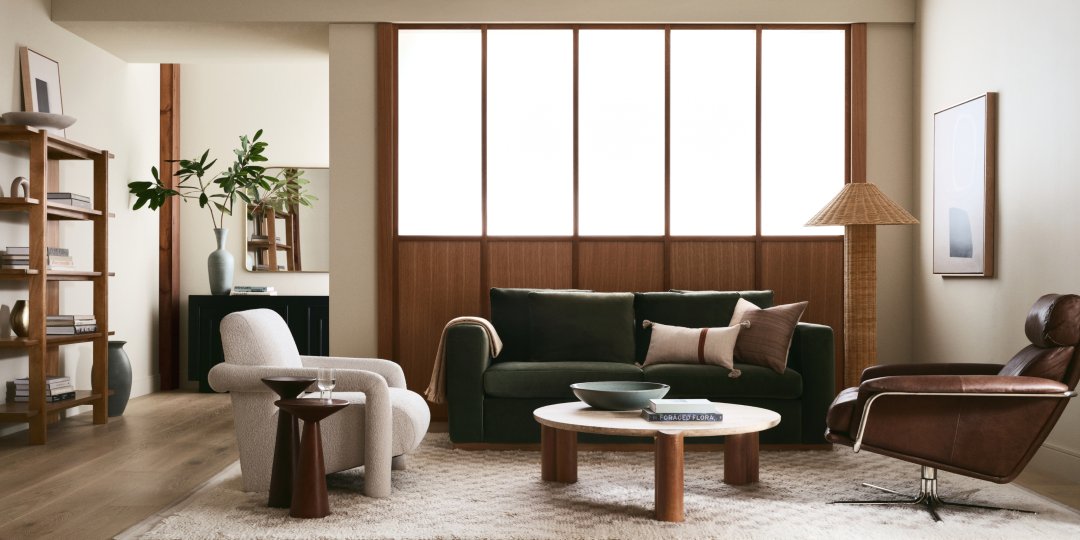 A modern, well-lit living room featuring a green velvet sofa, a white upholstered armchair, a brown leather recliner, a round wooden coffee table, and a beige textured rug. A floor lamp, bookshelf, and large framed window complete the cozy, minimalist decor.