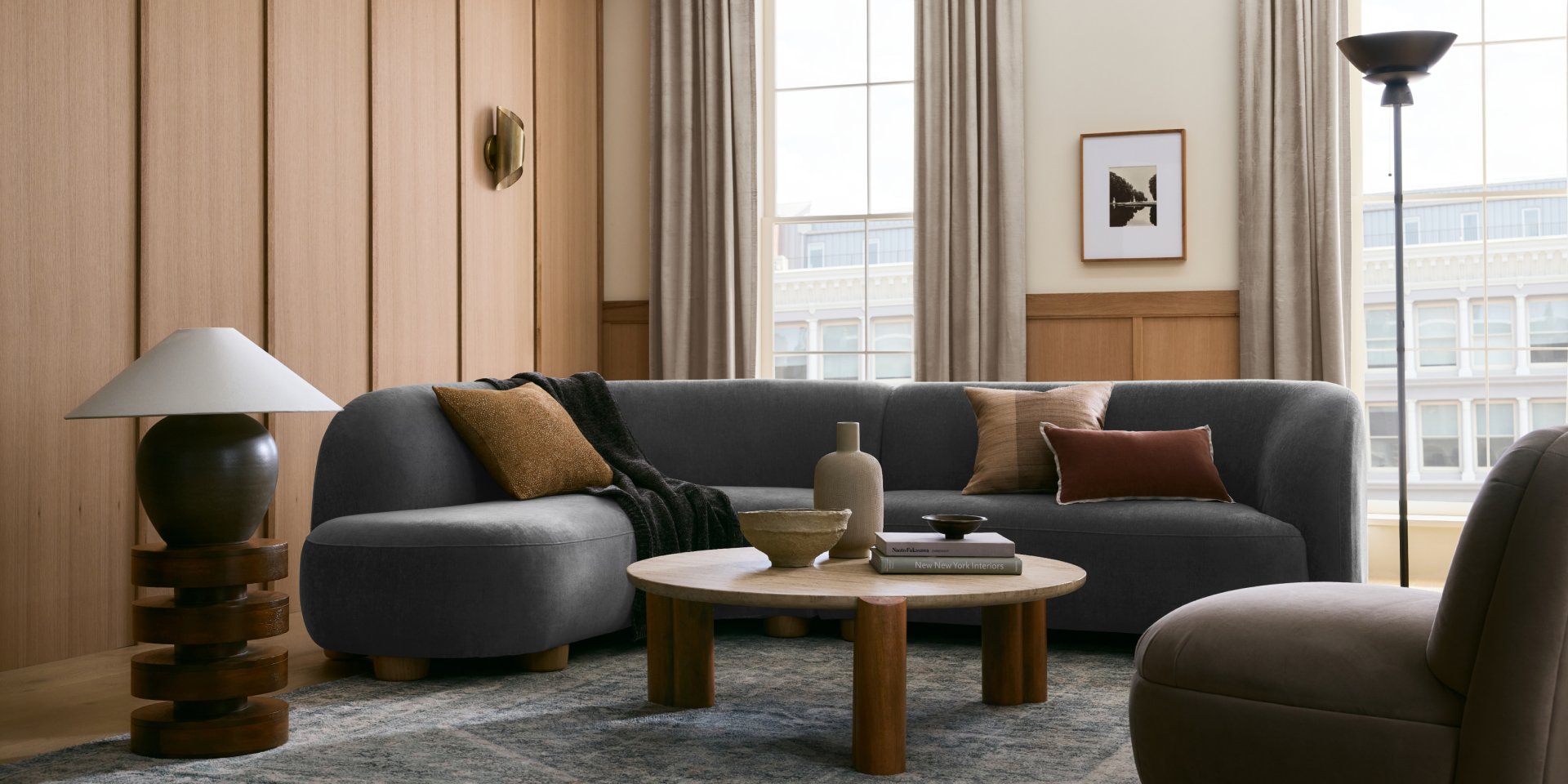 A modern living room features a grey L-shaped sofa with various pillows and a throw blanket. A round wooden coffee table holds decorative vases and a bowl. Large windows with curtains allow natural light to fill the room. A standing lamp is placed in the corner.