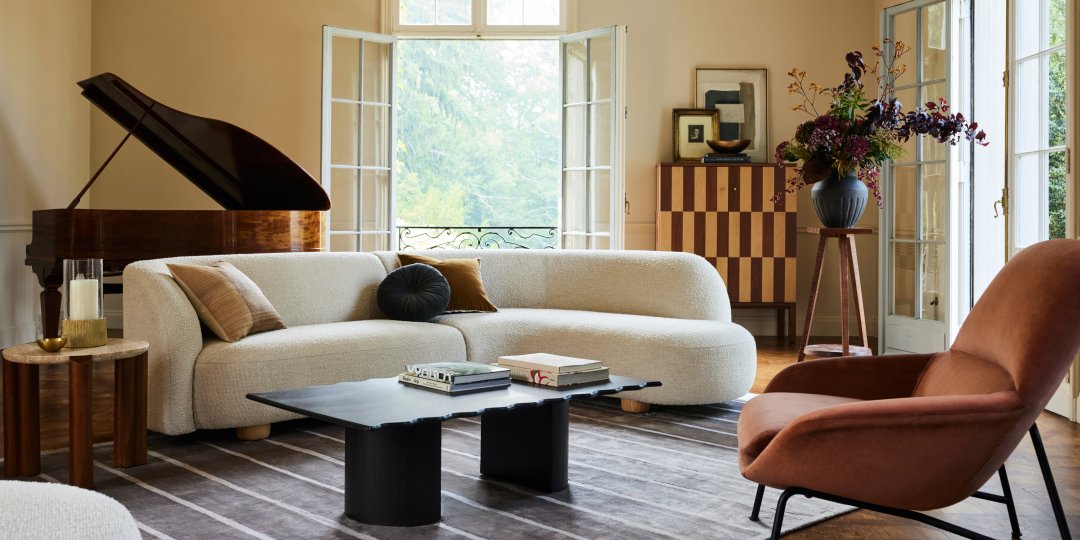 A stylish living room with a white curved sofa, brown armchair, black coffee table, and striped rug. A grand piano sits in the corner near wide-open French doors, and a checkered wooden cabinet with a flower arrangement stands against the wall.