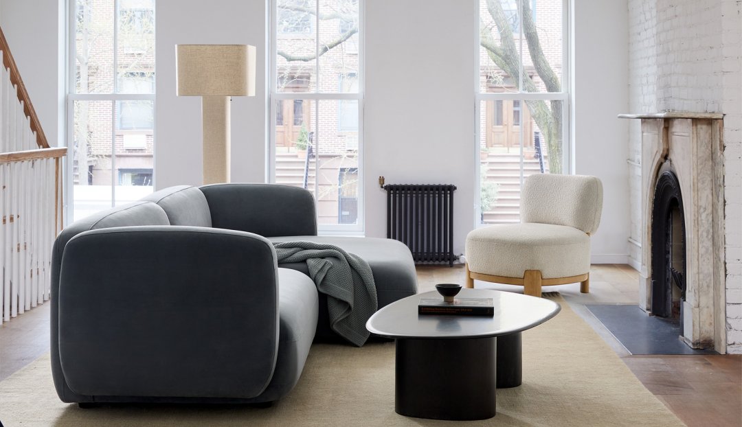 A modern living room features a curved gray sofa with a gray blanket, a rounded white chair, a black coffee table with books and a bowl, a tall standing lamp, and a fireplace. Large windows provide natural light, showing tree branches outside.