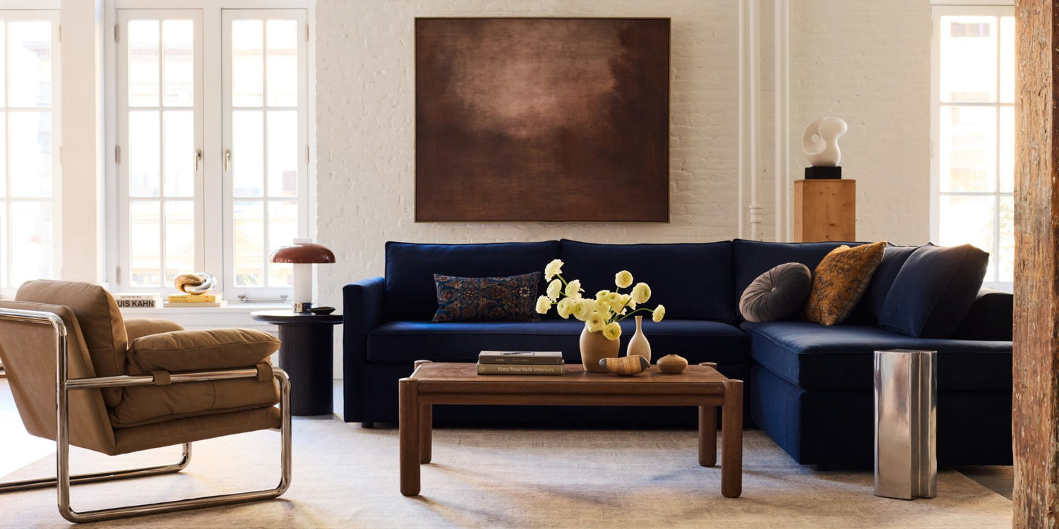 A modern living room with a navy blue sectional sofa, beige armchair, and wooden coffee table. The table has a vase of yellow flowers and books. Large windows and a minimalist brown painting on a white brick wall provide a bright, airy atmosphere.