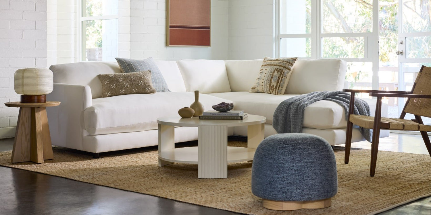A cozy living room featuring a white sectional sofa with various pillows, a round coffee table with decorative items, a wooden side table with a lamp, a woven indoor chair, a gray ottoman, and a large rug. The room is bright with natural light from large windows.