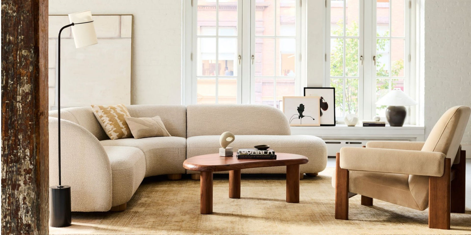 A modern living room with a cream-colored sectional sofa, a wooden coffee table, and an armchair with a beige cushion. A black floor lamp stands to the left. Large windows with white frames let in natural light, illuminating minimalist decor and wall art.