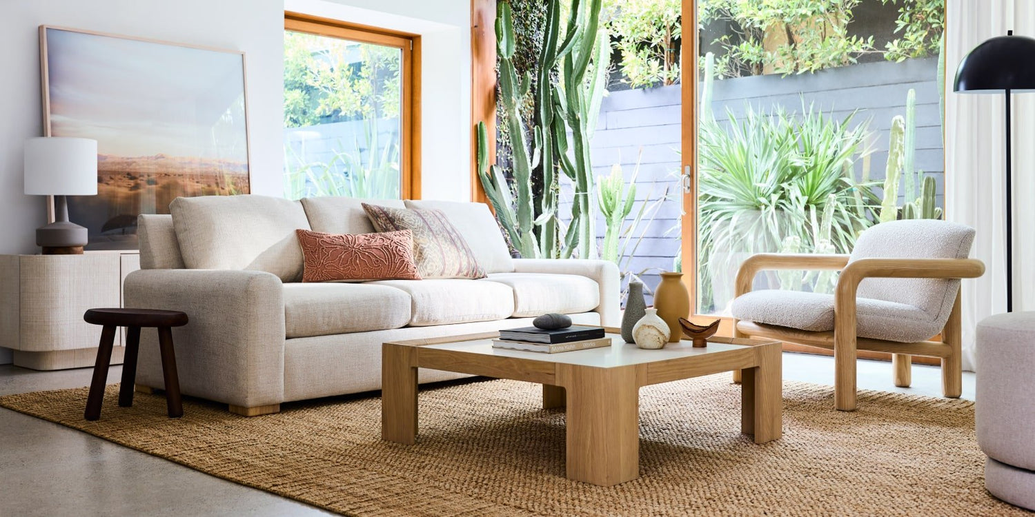 A modern living room with a beige sofa and a matching armchair. The room features a wooden coffee table, a side table with a lamp, a large framed landscape picture, and floor-to-ceiling windows showcasing green plants and natural light.