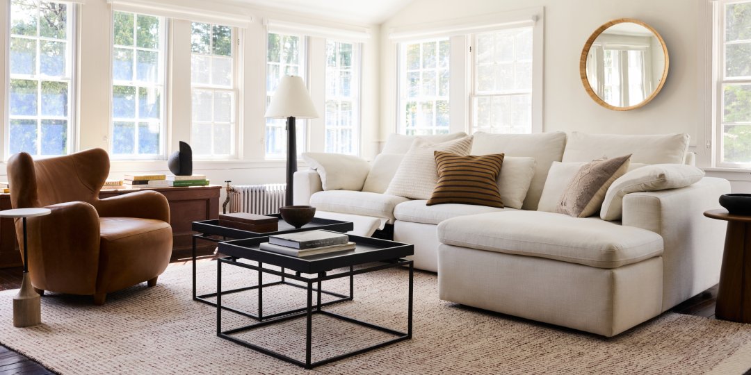 A bright living room with large windows and white walls. It features a cream sectional sofa with assorted pillows, a brown leather armchair, and a black coffee table on a textured rug. A round mirror hangs on the wall, and various decor items are placed on surfaces.