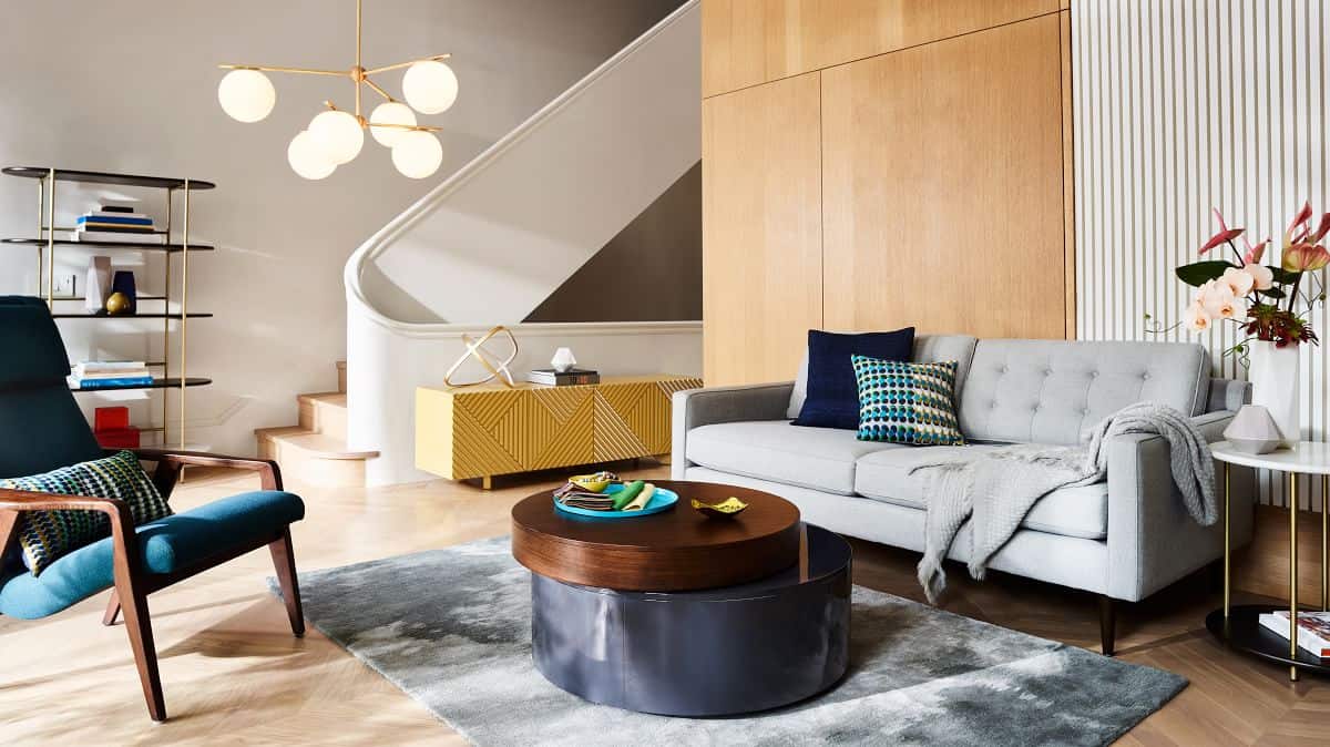 A modern living room featuring a light gray tufted sofa with colorful throw pillows, a round wooden coffee table on a gray rug, a dark green armchair, a contemporary chandelier, and a yellow sideboard. The room has a staircase with a white railing and wooden accents.