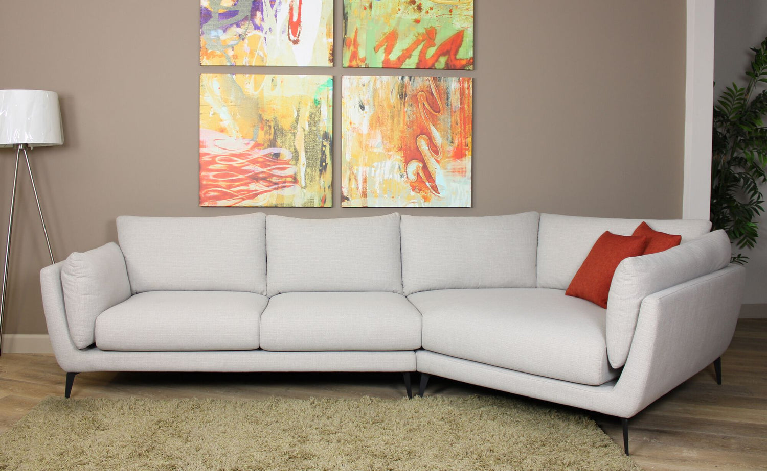 A stylish living room with a light grey sectional sofa adorned with a red accent pillow. The sofa is placed on a green rug, beneath a set of vibrant abstract paintings on a taupe wall. A white floor lamp stands to the left and a potted plant is on the right.