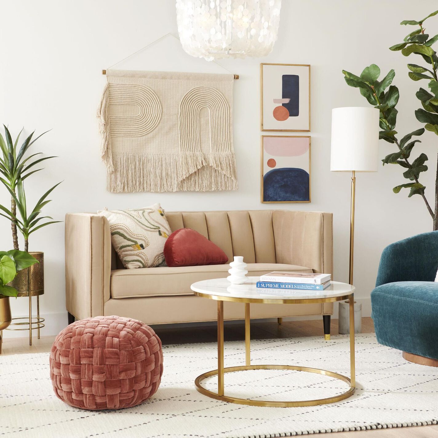 A modern living room features a beige sofa with a red cushion and a decorative pillow, a woven wall hanging above, and two abstract framed artworks. A green armchair, a marble coffee table with gold legs, a round textured red ottoman, and plants complete the space.