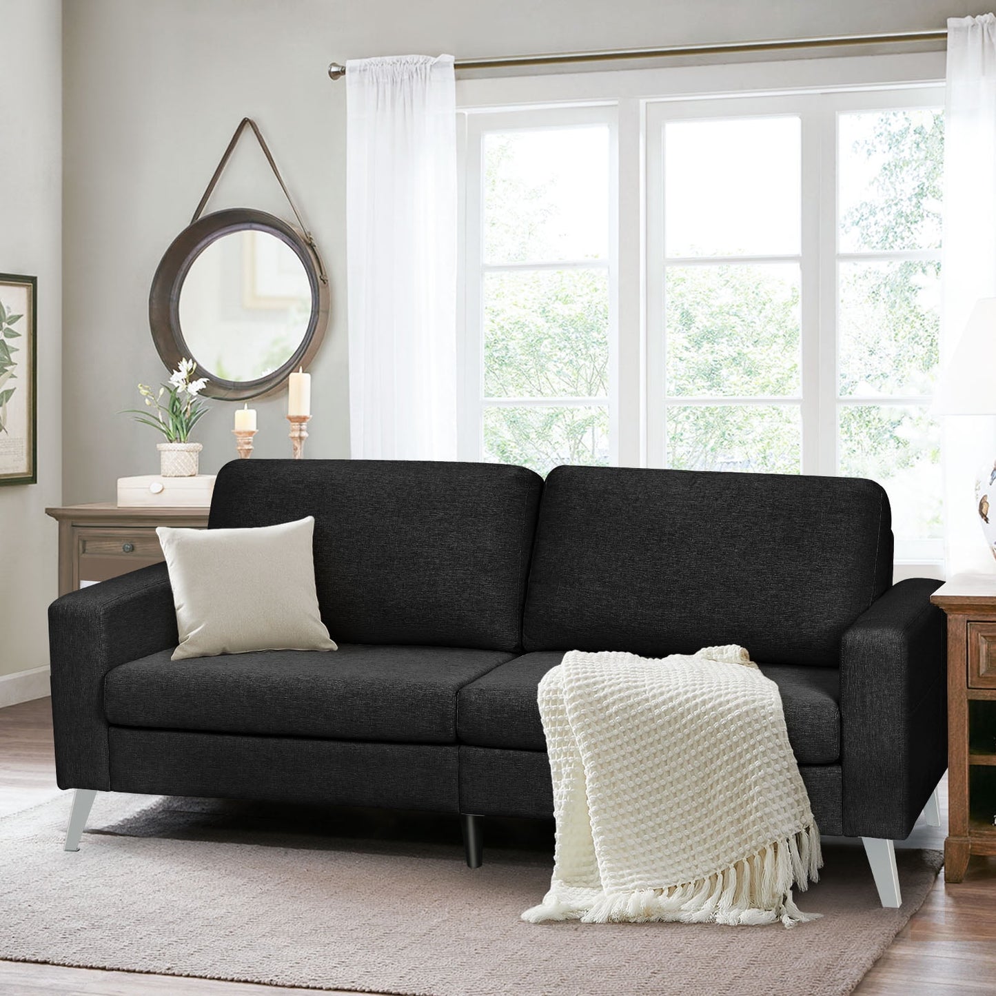 In a cozy living room, the YODOLLA 79" Sectional Sofa in dark gray is adorned with white cushions and a knit blanket. The space is enhanced by a round mirror, potted plants, candles, and a large window with sheer curtains that allow natural light to fill the room.