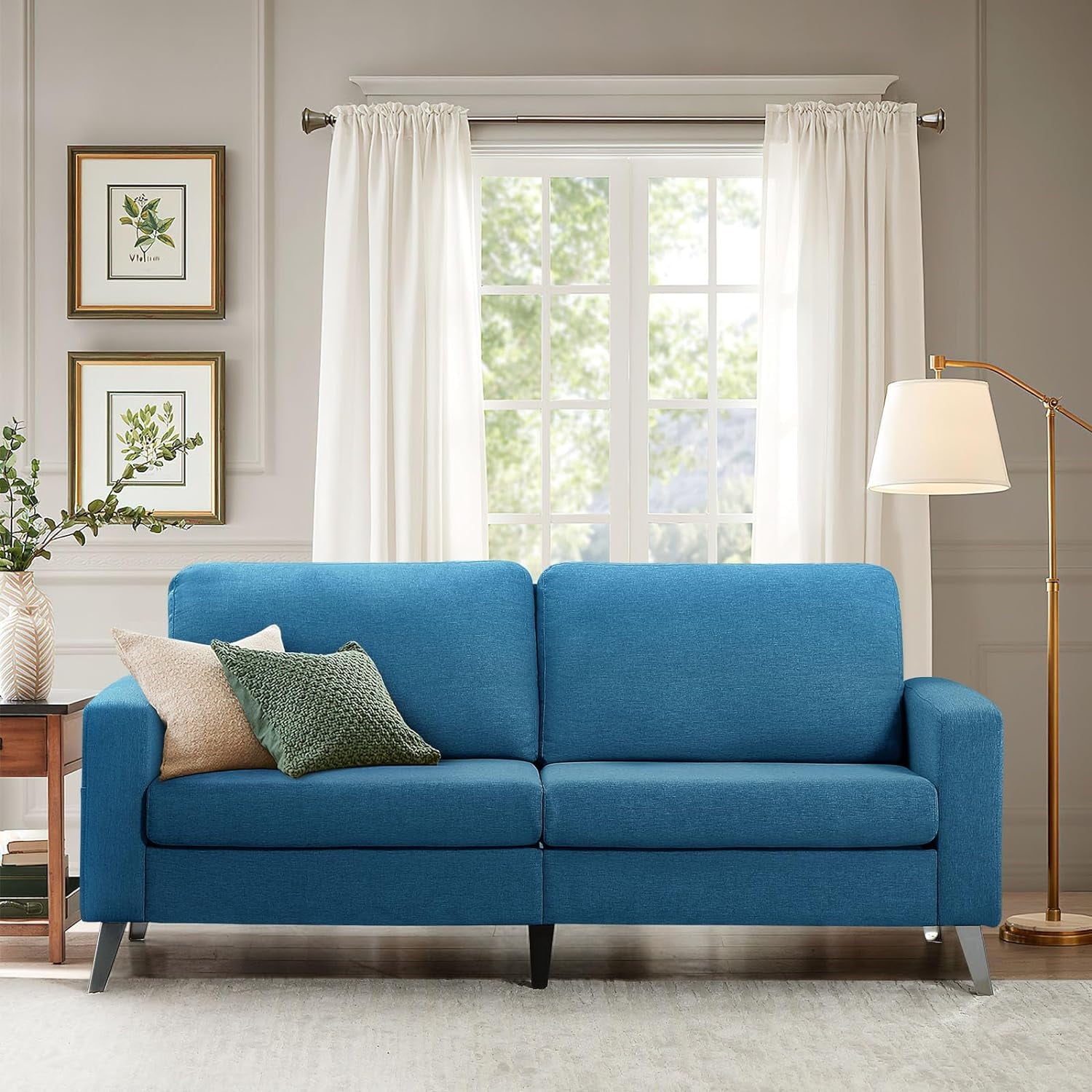 A stylish living room features the YODOLLA 79" Sectional Sofa adorned with green and beige pillows. A floor lamp stands nearby, while two framed botanical prints decorate the wall. Sunlight filters through a large window with white curtains, casting a serene glow.