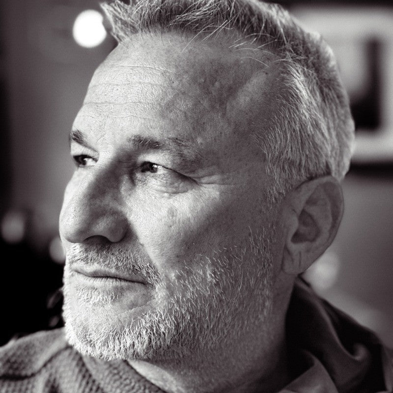 Black and white close-up portrait of an older man with short gray hair and a beard, looking thoughtfully into the distance. The background is blurred.