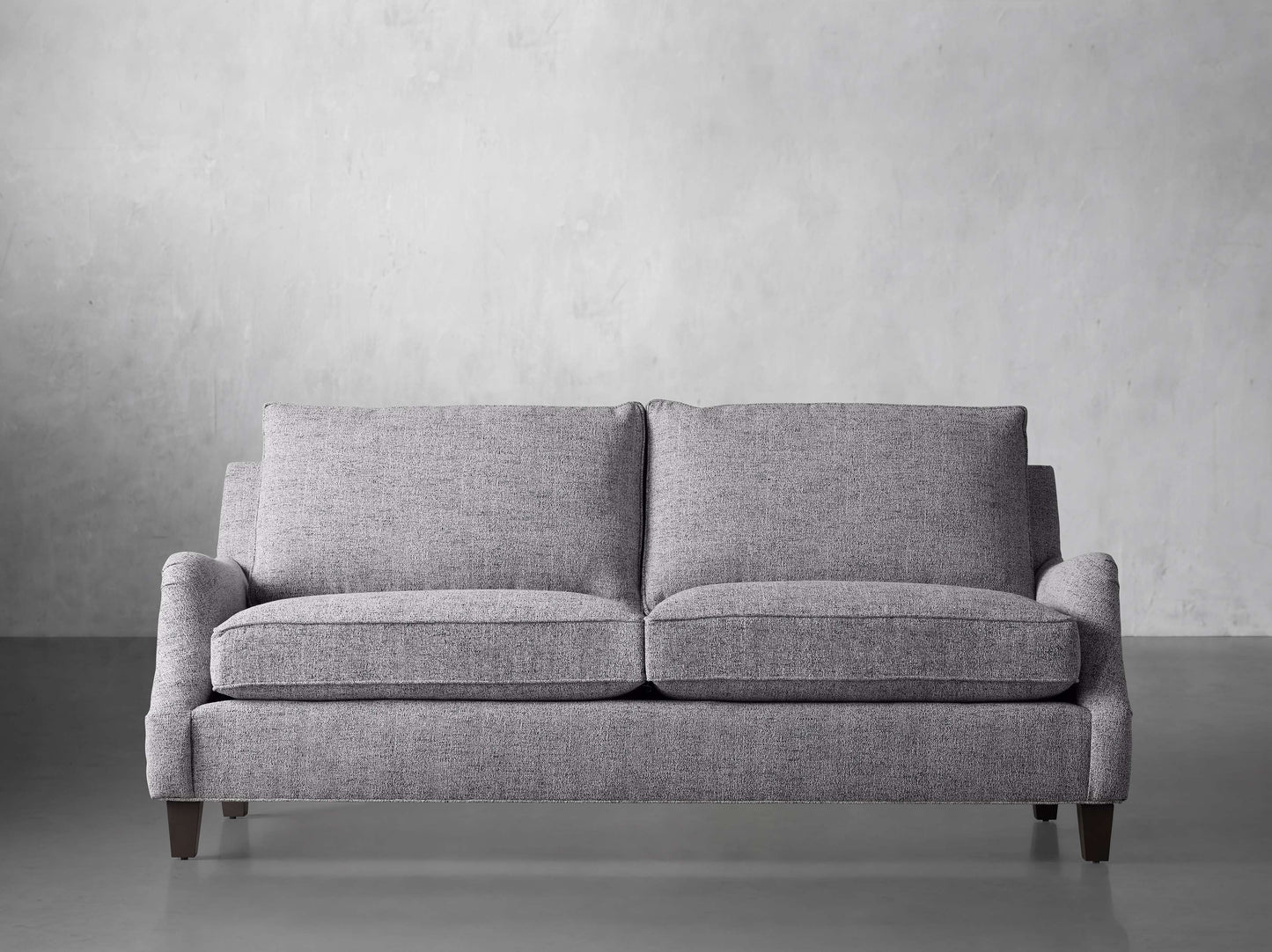 The Paxton 82" Sofa, featuring a modern gray design, is situated in front of a light gray textured wall. Its fabric looks soft, and it is supported by dark wooden legs.