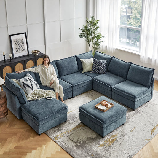 A woman relaxes on the spacious Victoria Oversized Soft Sectional Sofa with Storage in a bright living room. The decor includes a patterned rug, several cushions, a potted plant, and a sideboard adorned with various decorative items. Sunlight streams through large windows that are dressed with sheer curtains.