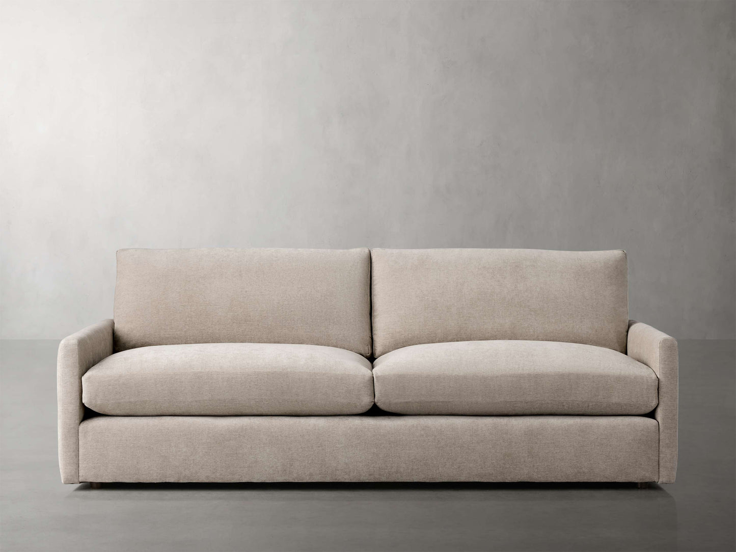 The Kipton 84" Deep Sofa, with its modern design and light beige color, features straight, clean lines set against a minimalist gray wall background. It has a plush appearance with two large seat and back cushions, resting on a smooth, polished floor.