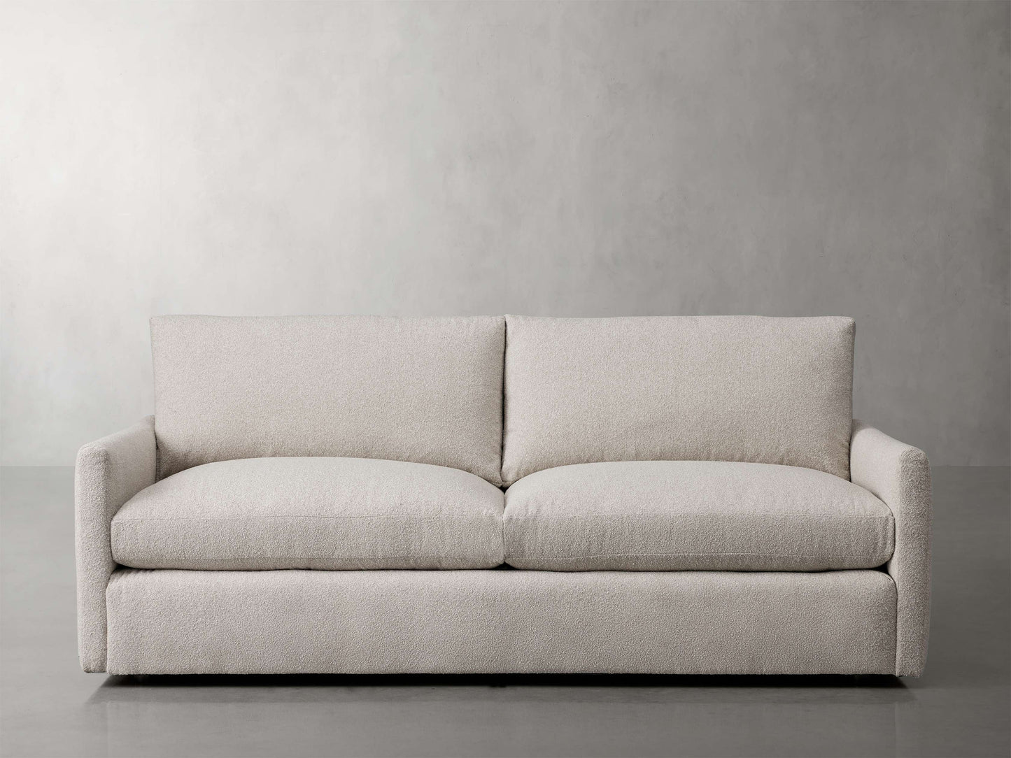 The Kipton 84" Deep Sofa, featuring a modern beige fabric with clean lines and plush cushions, is centered against a light grey textured wall on a smooth floor. Its wide armrests and minimalist design contribute to creating a cozy yet elegant living room atmosphere.