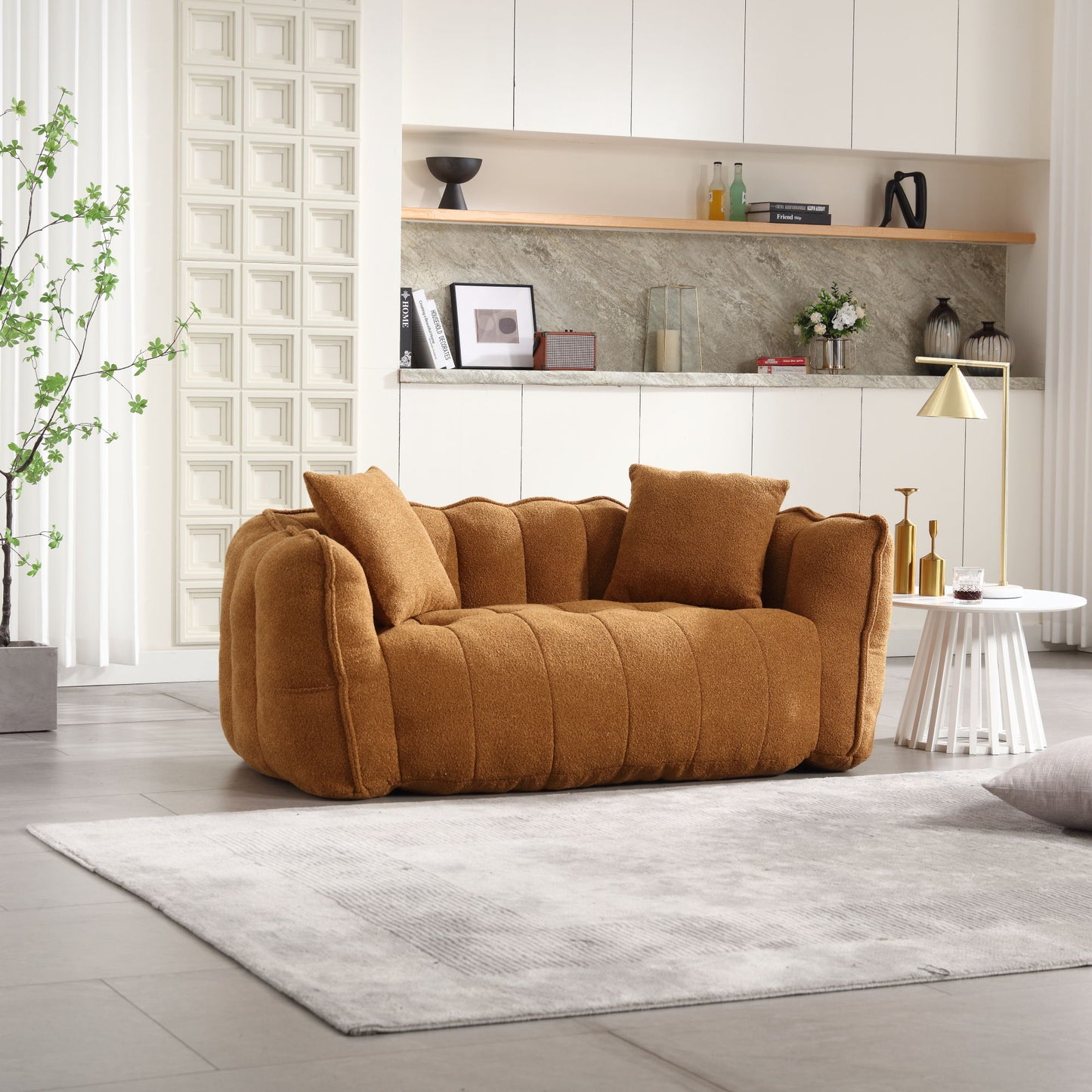 A modern living room includes a plush brown beanbag chair for two, set on a light gray rug. The background showcases white cabinets, a minimalist shelf with decor items, and a potted plant, creating a cozy and contemporary atmosphere.