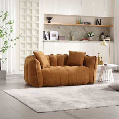 A modern living room includes a plush brown beanbag chair for two, set on a light gray rug. The background showcases white cabinets, a minimalist shelf with decor items, and a potted plant, creating a cozy and contemporary atmosphere.