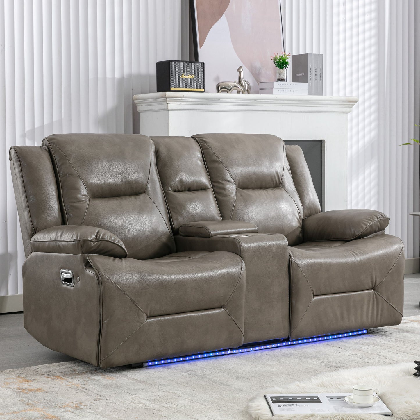 In a living room, the 2-Seater Home Theater Recliner features gray leather upholstery with manual reclining, two cup holders, and a storage box. Blue LED lights illuminate its base. A white fireplace with decor stands behind it, while a magazine rests on a light-colored rug.