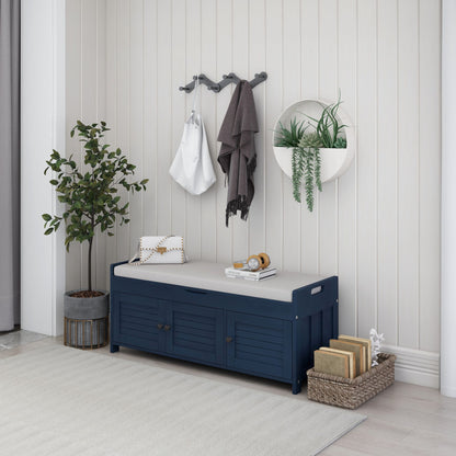 An organized entryway features a navy storage bench with shutter-shaped doors, topped with a tray and small plant. Hooks above hold bags and a scarf, next to a round shelf of hanging plants. A basket of books and tall plant enhance the light-paneled wall decor.