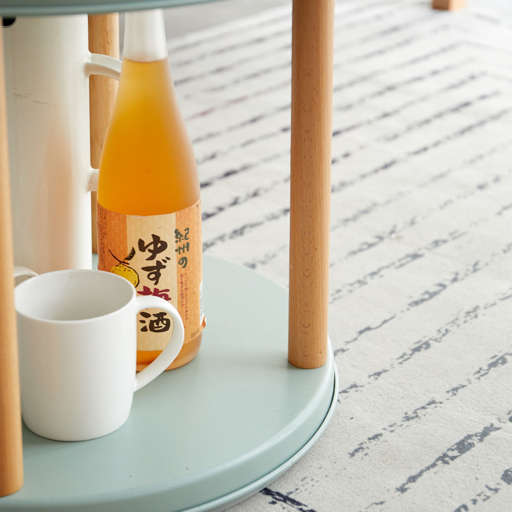 A Japanese yuzu-flavored beverage and a plain white mug sit on a light blue table with wooden legs, against a backdrop of a white rug with dark blue stripes. Nearby is the Storage Shelf, 360° Rotating Bookshelf, Bookcase with Large-Capacity Storage Space—ideal for any living room.