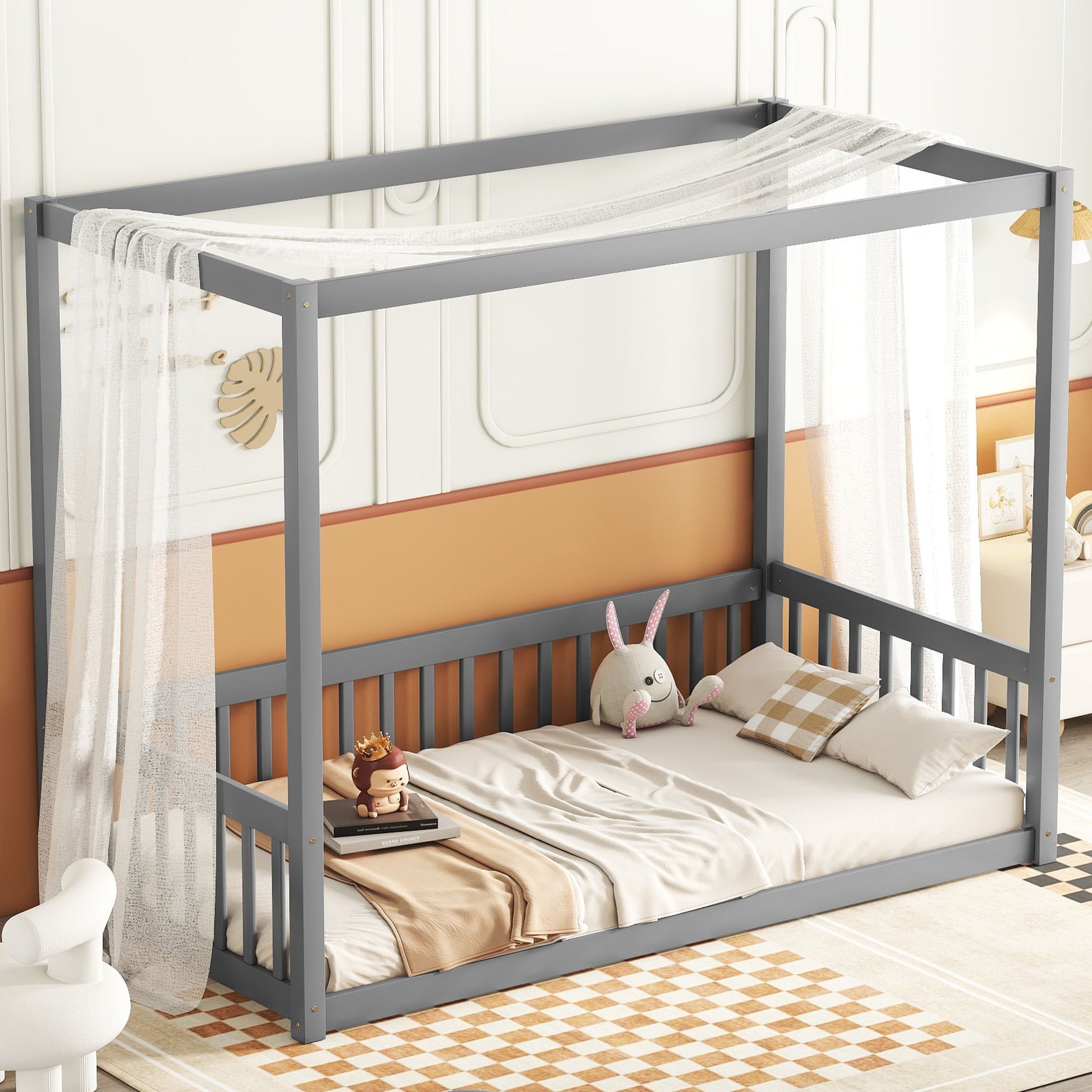 The product is a Canopy Frame Floor Bed With Fence, Guardrails, featuring a gray frame with sheer white curtains, beige and white checkered blanket and two pillows. A plush rabbit and toy lion sit on the bed. The floor sports a checkered rug, while the wall is painted light peach and white.