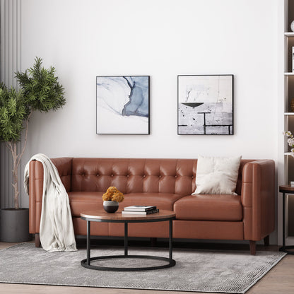 A stylish living room features the "Comfy 3 Seat Sofa With Tufted Back" in brown leather, adorned with a white pillow and throw. Two abstract paintings hang on the white wall above. A round coffee table holds a plant centerpiece in front, while a tall potted plant stands nearby.