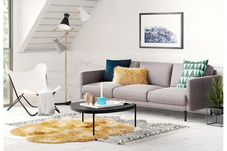 A modern living room features a gray sofa with teal and yellow pillows, a black and white patterned rug, and a round black coffee table. A fluffy yellow rug is beneath the table. A white chair, a floor lamp, and a framed abstract artwork adorn the space.