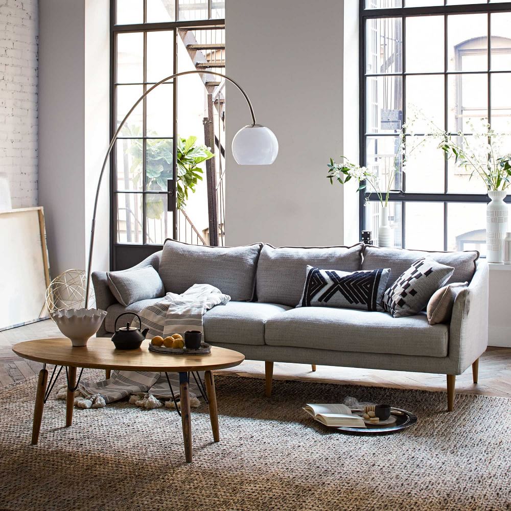 A modern living room with a large window, light gray sofa, and round wooden coffee table. The sofa has various cushions and a blanket. There's a tall, arched floor lamp and decor items including plants, books, and a bowl on the table. The room is bright and airy.