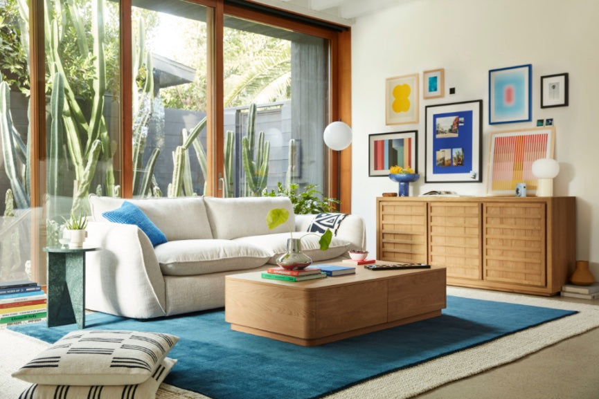 A modern living room with a beige sofa adorned with colorful cushions, a wooden coffee table on a blue rug, and a wooden cabinet against the wall. The wall is decorated with a variety of framed art pieces. A large window overlooks a garden with tall cacti.