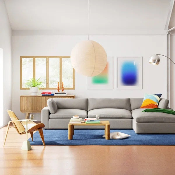 A modern living room with a gray sectional sofa adorned with colorful pillows, a wooden chair with a small side table, and a wooden coffee table atop a blue rug. Large windows allow natural light, and abstract art pieces hang on the wall. A spherical light fixture hangs from the ceiling.