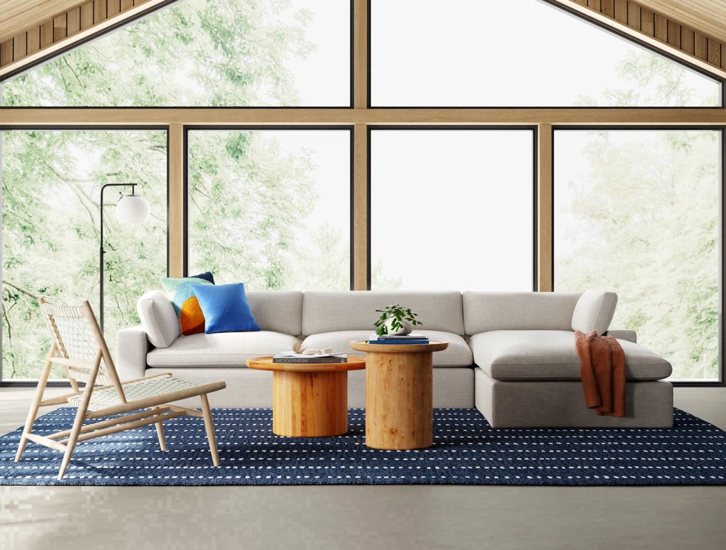 A modern living room with large windows revealing a lush, green forest outside. The room features a light grey sectional sofa adorned with colorful pillows, two wooden coffee tables, a woven chair, an orange throw blanket, a floor lamp, and a blue patterned rug.