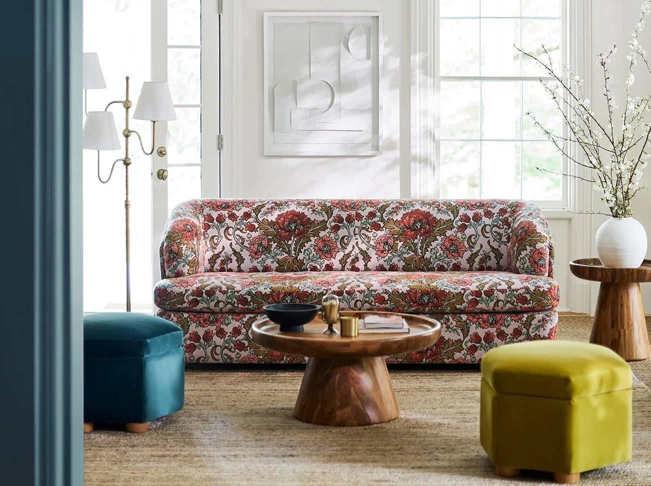 A brightly lit living room featuring a floral-patterned sofa, a wooden coffee table with a black bowl and decor, a blue ottoman, a yellow ottoman, a tall floor lamp, and a framed abstract artwork on a white wall. The space has several large windows and a light, airy feel.