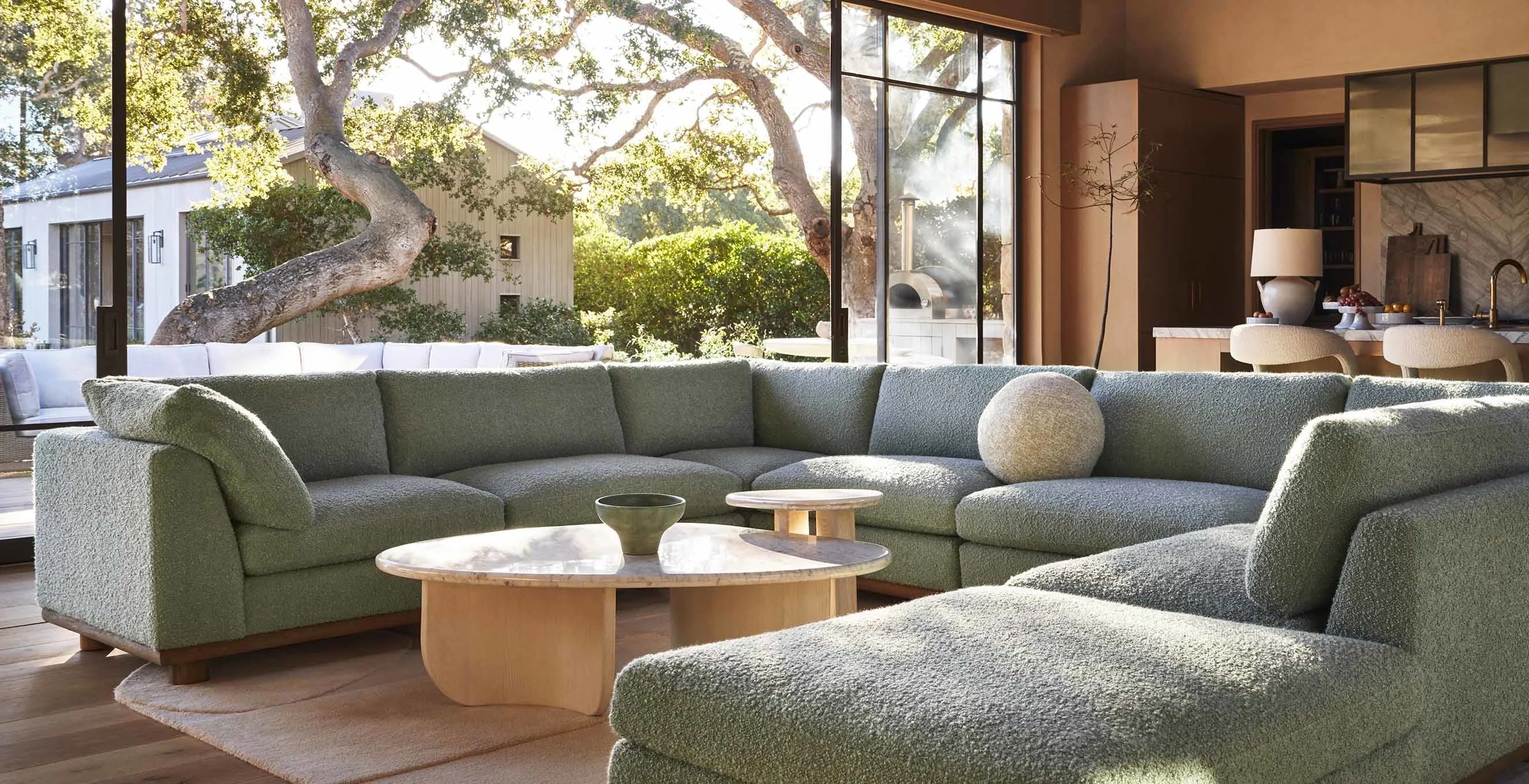 A spacious living room features a large, green sectional sofa arranged in an L-shape, centered around round, light-colored coffee tables. Large windows offer an unobstructed view of a lush, sunny outdoor area with trees and a building in the background.