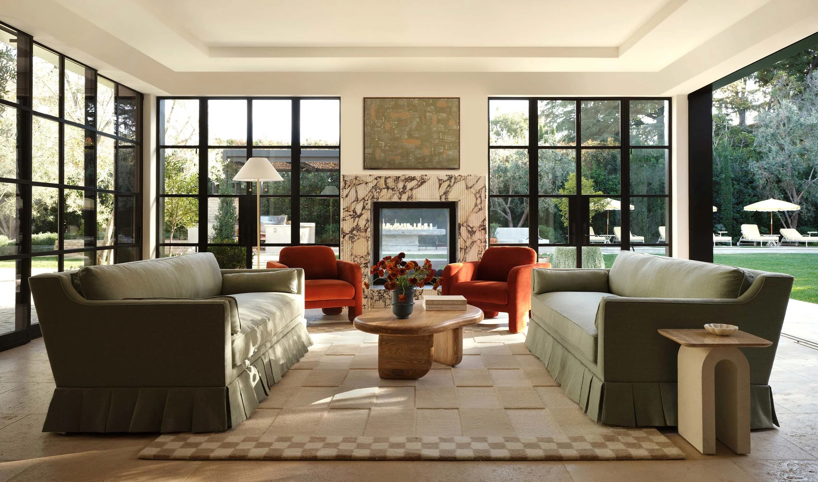 A modern, sunlit living room featuring two green couches and two red armchairs arranged around a wooden coffee table with a vase of flowers. The room has large windows, a marble fireplace, a contemporary rug, and opens up to a lush outdoor garden area.