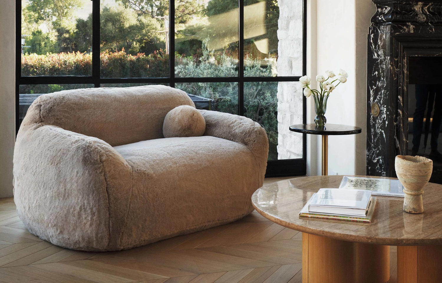 A cozy, plush beige sofa with a matching round cushion sits in a stylish living room with a large window overlooking greenery. A round wooden coffee table with books and a vase, and a small side table with white flowers, complete the scene.