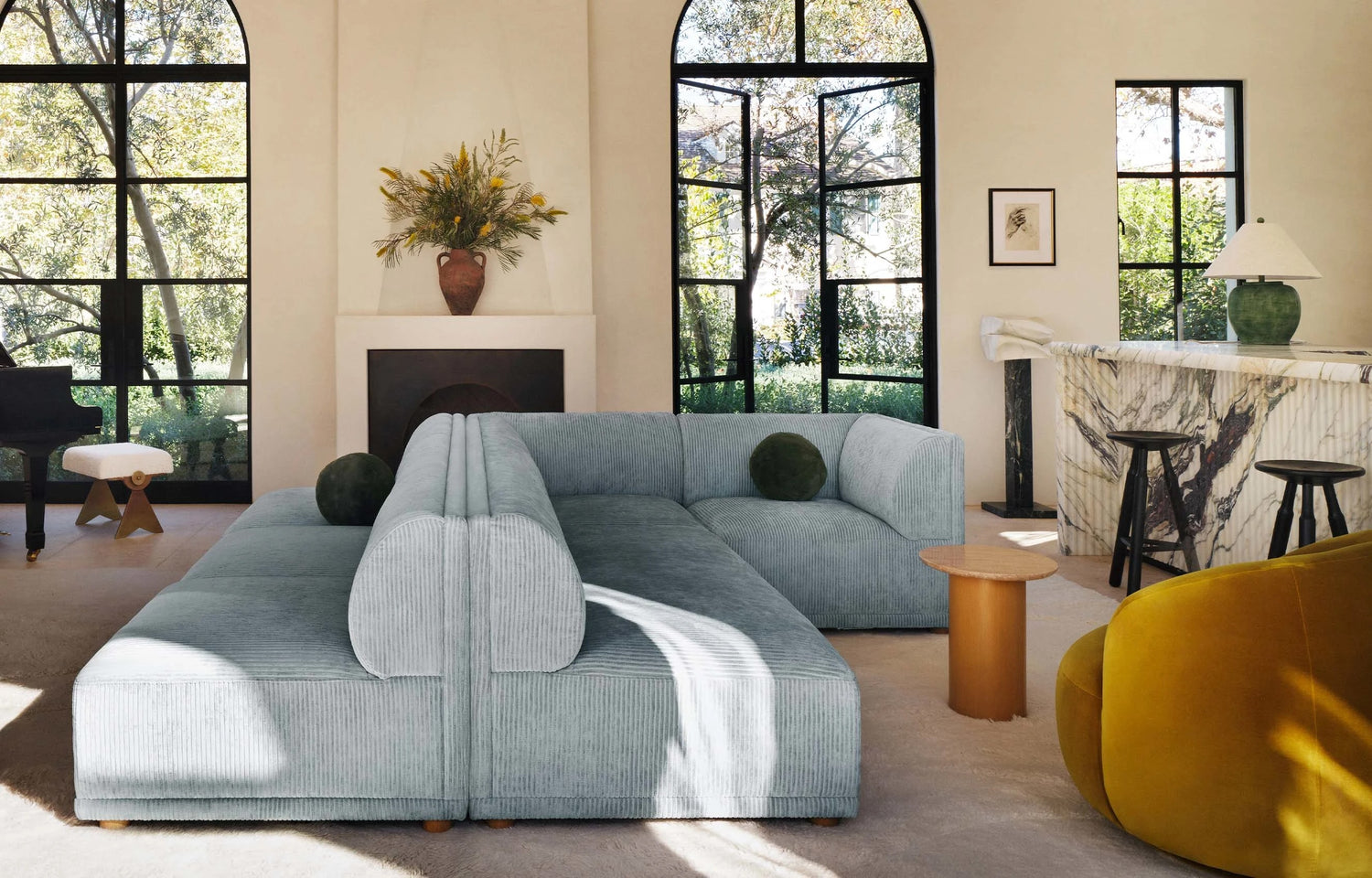 A modern living room featuring a light blue sectional sofa, a yellow armchair, a coffee table, and a marble-topped bar area with high stools. Large arched windows offer a view of lush greenery, and sunlight streams in, illuminating the space. A vase with flowers is on the mantel.
