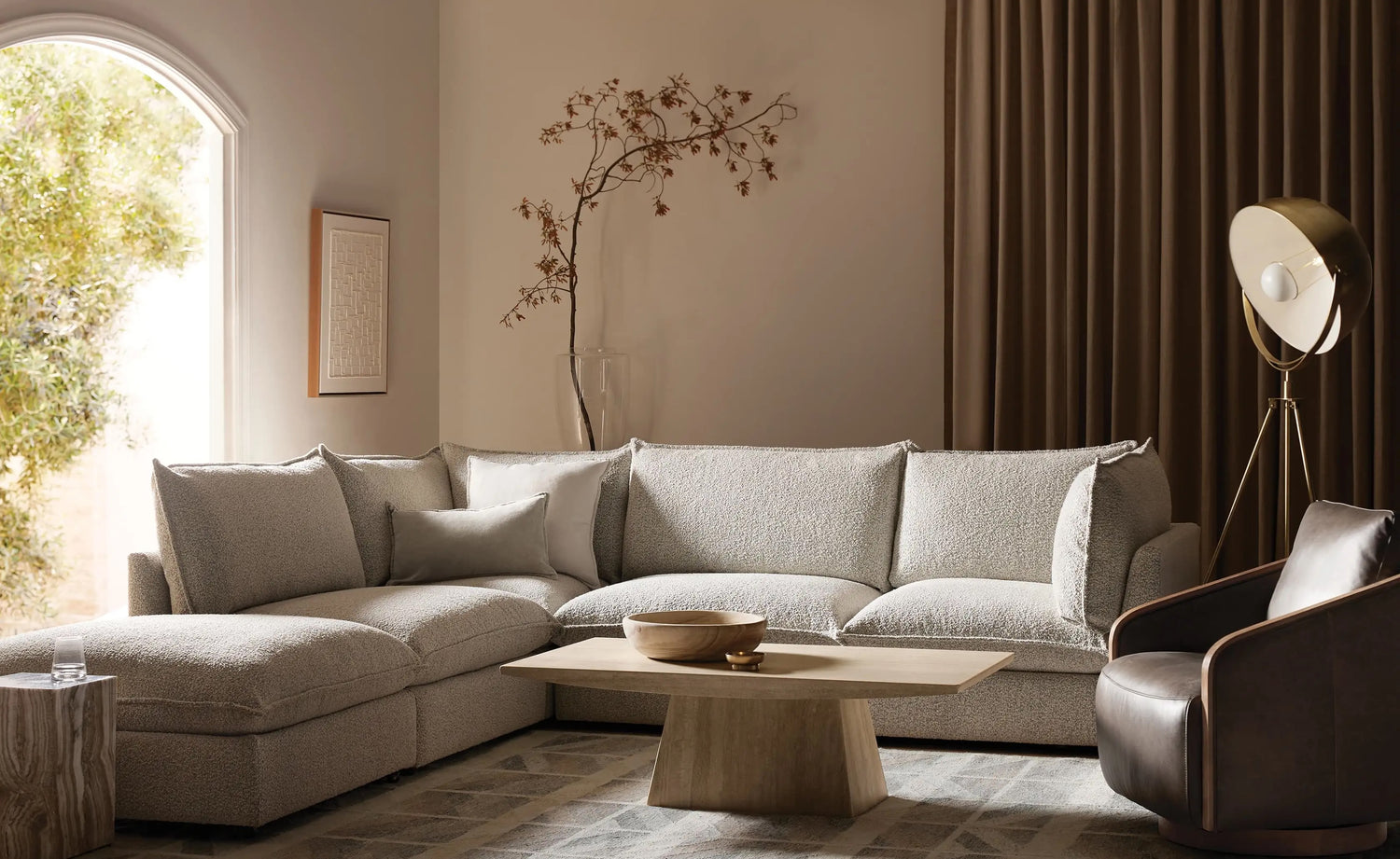 A modern living room featuring a light gray sectional sofa, a wooden coffee table with a bowl, and a brown leather armchair. The room is decorated with a large floor lamp, a piece of wall art, and a tall vase with dried branches, with natural light streaming through an arched window.