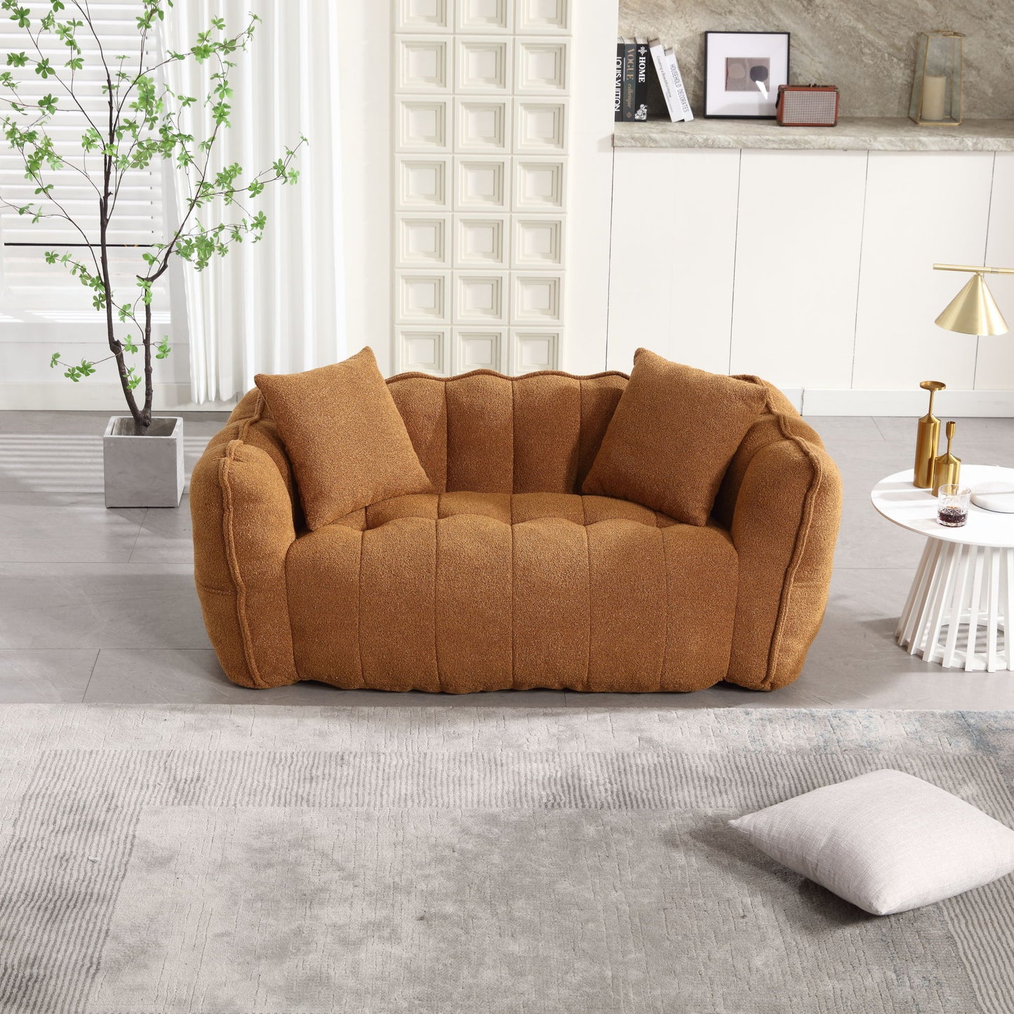 In a modern living room, a comfy brown sofa with tufting and two matching pillows complements a round white side table adorned with decor, a gray textured rug, and cream cabinets in the backdrop. Nearby sits the Soft Beanbag Chair with High Resilience Foam Core for Two People.