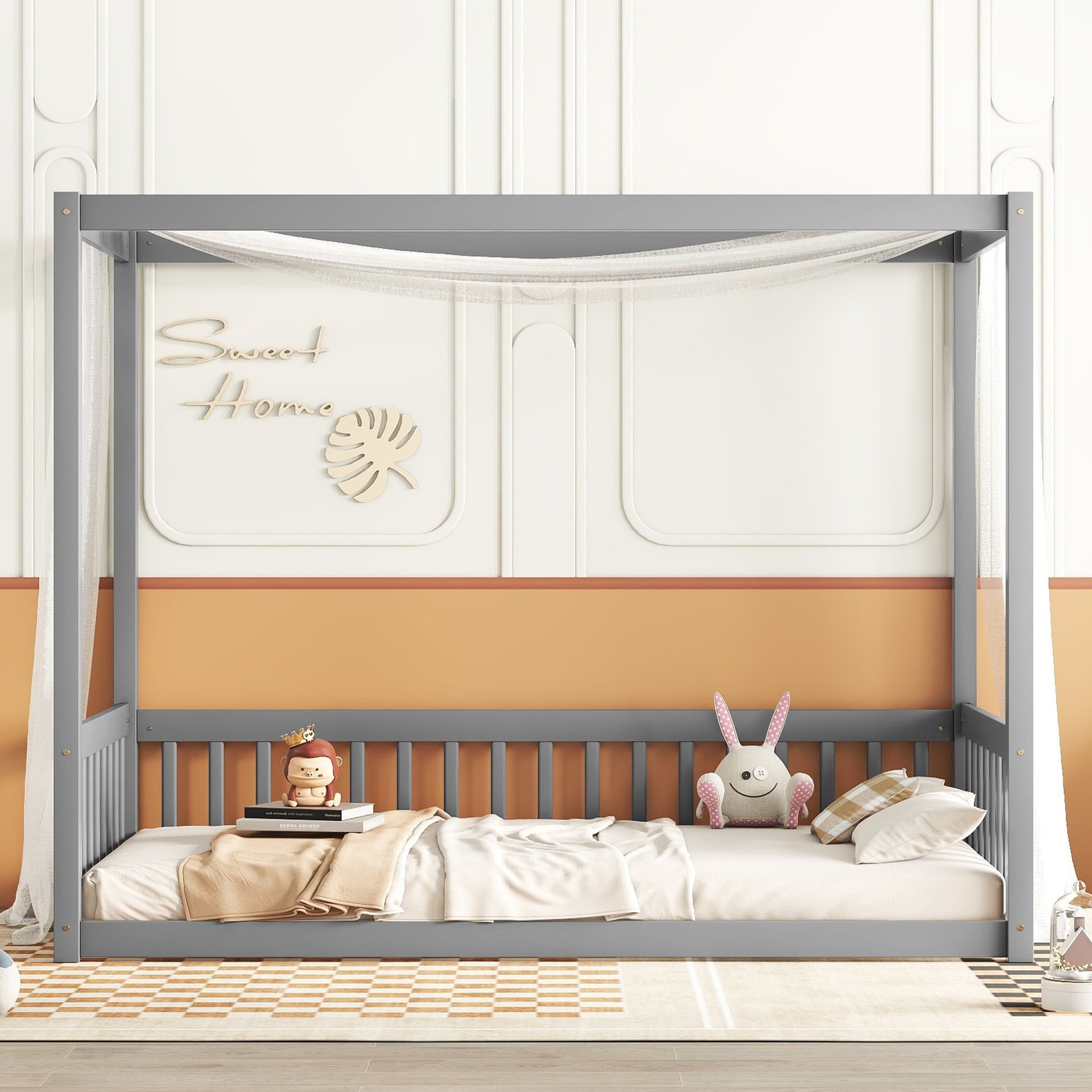 In a room, a Canopy Frame Floor Bed With Fence and Guardrails featuring a gray frame and white canopy stands decorated with a rabbit and doll. The peach-and-white wall displays "Sweet Home" with leafy decor, complemented by a checkered rug on the floor.